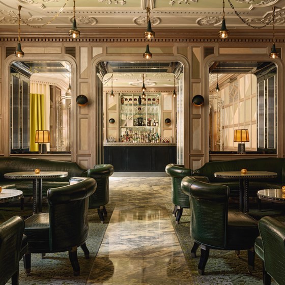 Elegant interior of The Connaught Bar, featuring deep green leather seating, mirrored wall panels, intricate ceiling detailing, and a softly lit central bar framed by arched woodwork.